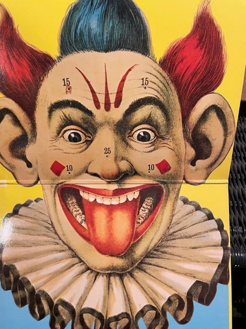 May include: A vintage carnival game target featuring a clown with a wide, toothy grin and a red tongue sticking out. The clown has red hair, blue hair, and a white ruffle collar. The target has numbers 10, 15, and 25 printed on it.