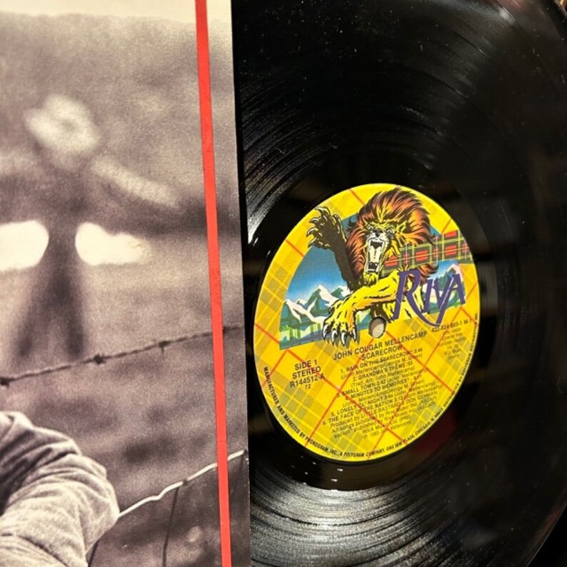 May include: A black vinyl record with a yellow and red label. The label features a lion with a red and black plaid background. The text on the label reads "John Cougar Mellencamp Scarecrow" and lists the songs on the record.