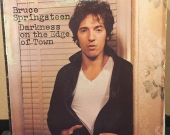 May include: A vinyl record album cover featuring Bruce Springsteen. The album title is "Darkness on the Edge of Town". The cover art features a close-up of Springsteen in a black leather jacket and white t-shirt, standing in front of a window with blinds.