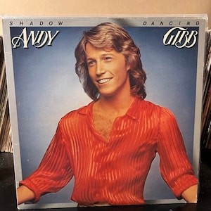 May include: A vintage vinyl record album cover featuring a portrait of Andy Gibb, a member of the Bee Gees, with the title "Shadow Dancing" printed in white on a blue background.