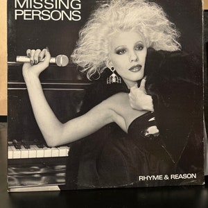 May include: A black and white photo of a woman with blonde hair holding a microphone. The photo is on a record album cover with the text "MISSING PERSONS" and "RHYME & REASON" on it.