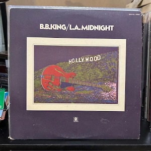 May include: Album cover for B.B. King's "L.A. Midnight." The cover is dark purple with the title in white. A framed image features a red guitar superimposed over a green and blue landscape with the word "HOLLYWOOD" above it.