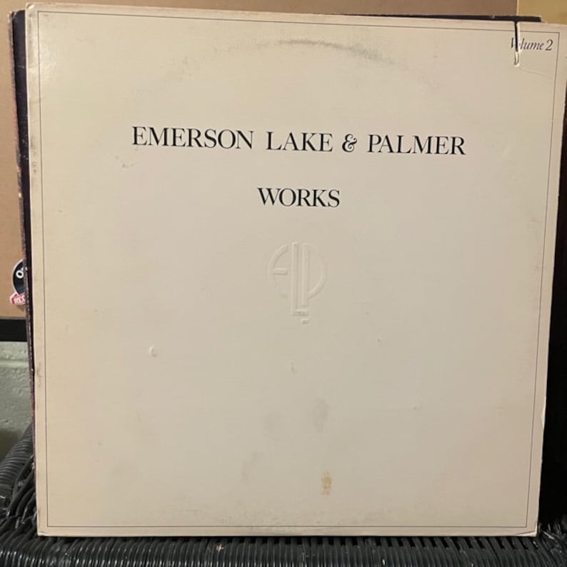 May include: A white vinyl record album cover with the text "EMERSON LAKE & PALMER" and "WORKS" in black lettering. The album cover also features the text "Volume 2" in the top right corner and a stylized "ELP" logo in the center.