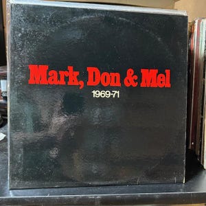 May include: A vinyl record album with a black cover. The album title "Mark, Don & Mel" is in red block letters, with "1969-71" below. The album is part of a collection of records.