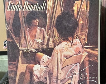 Könnte beinhalten: Ein Vintage-Vinyl-Schallplattencover mit Linda Ronstadt. Das Cover zeigt Ronstadt in einem weißen Gewand, auf einem Stuhl sitzend, mit einem Spiegel, der ihr Bild reflektiert. Der Albumtitel "Simple Dreams" ist in Kursivschrift am unteren Rand des Covers geschrieben.