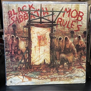 May include: A vinyl record cover featuring the Black Sabbath Mob Rules album artwork. The illustration depicts hooded figures in tattered robes standing before a wooden structure with a blood-stained cloth, set against a brick wall background.