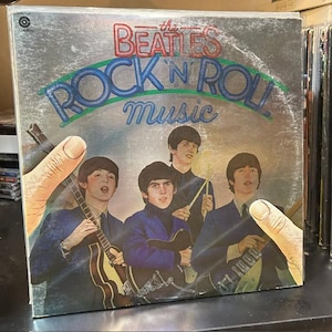 Pu&ograve; includere: La copertina del disco in vinile The Beatles Rock 'n' Roll Music presenta uno sfondo argentato con il nome della band in rosso e il titolo in blu. Quattro membri della band sono raffigurati in abiti blu, con due mani che incorniciano l'immagine ai lati.