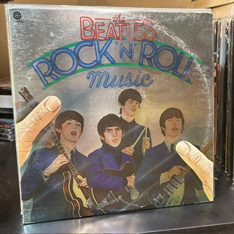 Pu&ograve; includere: La copertina del disco in vinile The Beatles Rock 'n' Roll Music presenta uno sfondo argentato con il nome della band in rosso e il titolo in blu. Quattro membri della band sono raffigurati in abiti blu, con due mani che incorniciano l'immagine ai lati.