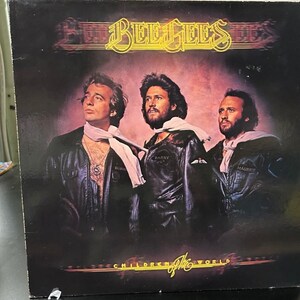 May include: A vintage vinyl record album cover featuring the Bee Gees. The cover art features a photo of the three brothers, Barry, Robin, and Maurice, in leather jackets and scarves. The album title, "Children of the World", is printed in gold lettering at the bottom of the cover.