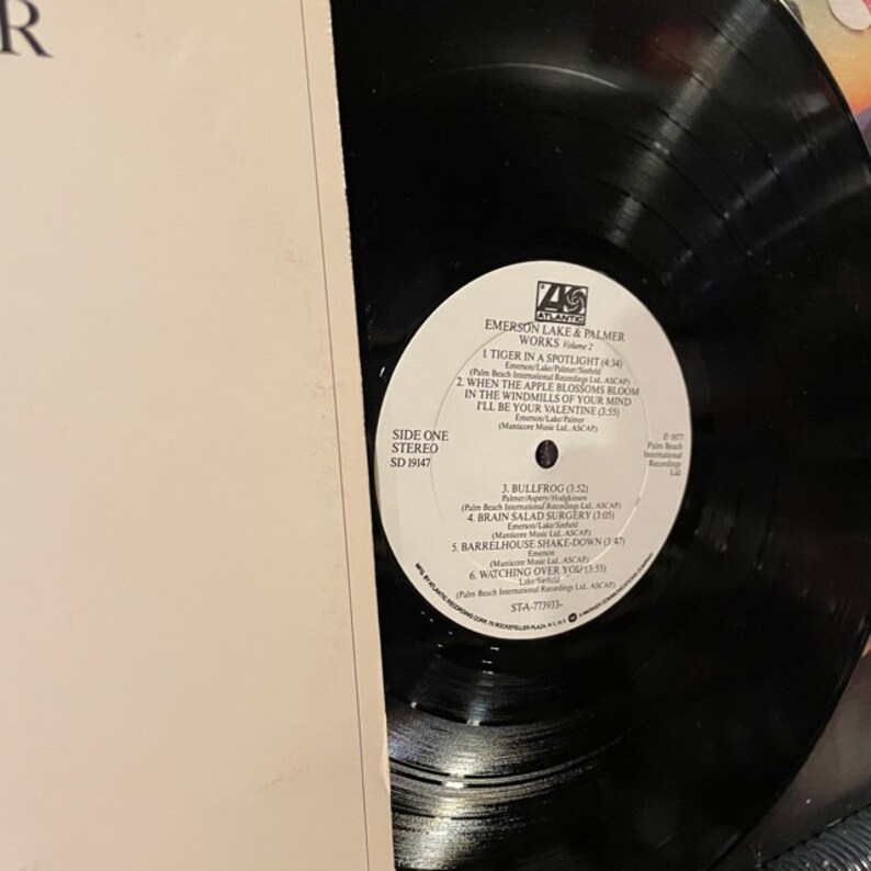 May include: A black vinyl record with a white label. The label has the text "Emerson Lake & Palmer Works Volume 1" and lists the songs "Tiger in a Spotlight", "When the Apple Blossoms Bloom in the Windmills of Your Mind I'll Be Your Valentine", "Bullfrog", "Brain Salad Surgery", "Barrelhouse Shake-Down", and "Watching Over You". The record is labeled "Side One Stereo SD 19147".