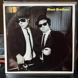 May include: Black and white photo of two men wearing suits and sunglasses. The men are standing in front of a striped wall. The text "Blues Brothers" is printed in yellow letters at the top of the photo.