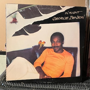 May include: A vinyl record album cover featuring a man in an orange shirt sitting on a couch with two yellow birds. The album title is "In Flight" by "George Benson".
