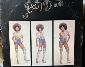 May include: A vintage album cover featuring the name "Betty Davis" in silver script. The cover displays three images of a woman in a floral top, denim shorts, and silver thigh-high boots. The background is black.