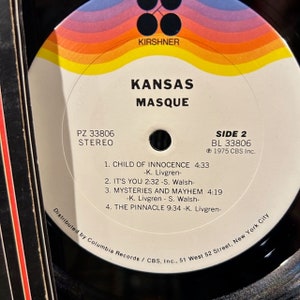 May include: A black vinyl record with a white label. The label has a red, yellow and orange design. The text on the label reads "Kansas Masque" and "Side 2 BL 33806 ℗ 1975 CBS Inc."