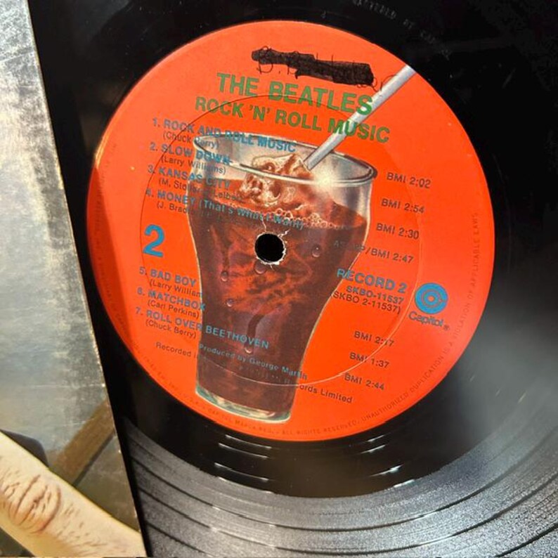 Pu&ograve; includere: Un'etichetta per disco in vinile di The Beatles Rock 'n' Roll Music, Record 2. L'etichetta &egrave; arancione brillante con testo verde e presenta una grafica centrale di un bicchiere pieno di una bevanda gassata scura con una cannuccia. La tracklist include Rock and Roll Music, Slow Down, Kansas City, Money, Bad Boy, Matchbox e Roll Over Beethoven.
