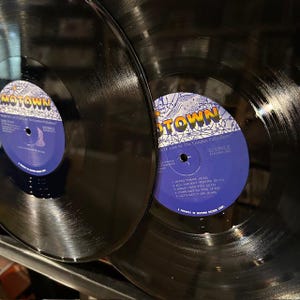 May include: Two black vinyl records with blue and white labels. The labels read "MOTOWN" and list song titles. The records are in good condition, with visible grooves and a glossy finish. The records are likely vintage.