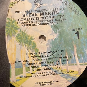 May include: A black and white vinyl record with a label featuring a photo of palm trees and the text "Home of Warner Bros. Records". The label also includes the text "William E. McEuen Presents Steve Martin Comedy Is Not Pretty" and "Produced by William E. McEuen Aspen Recording Society". The record is labeled "HS 3392" and includes a list of songs: "Born to Be Wild", "The All Being", "McDonald's/Men's Underwear", "Drop Thumb Medley", "Googlephonics", and "Hostages".