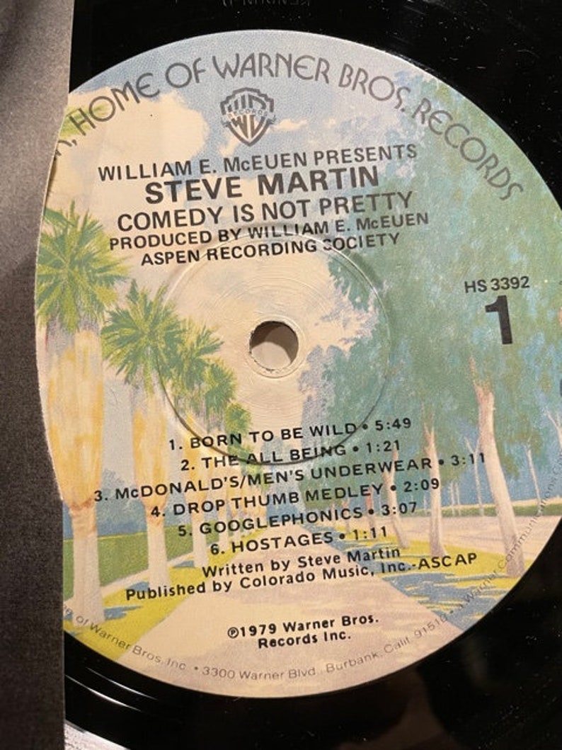 May include: A black and white vinyl record with a label featuring a photo of palm trees and the text "Home of Warner Bros. Records". The label also includes the text "William E. McEuen Presents Steve Martin Comedy Is Not Pretty" and "Produced by William E. McEuen Aspen Recording Society". The record is labeled "HS 3392" and includes a list of songs: "Born to Be Wild", "The All Being", "McDonald's/Men's Underwear", "Drop Thumb Medley", "Googlephonics", and "Hostages".