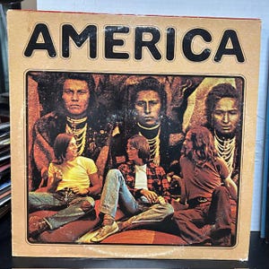 May include: Vintage vinyl record album cover for the band America. The cover features the band members sitting in front of a backdrop of Native American portraits. The album title "AMERICA" is printed in bold, black letters at the top.
