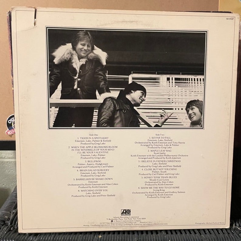 May include: Back cover of a vinyl record album featuring a black and white photo of three men. The album is titled "Emerson, Lake & Palmer" and features the song titles "Tiger in a Spotlight", "When the Apple Blossoms Bloom in the Windmills of Your Mind", "I'll Be Your Valentine", "Bullfrog", "Brain Salad Surgery", "Barrelhouse Shake Down", "Watching Over You", "So Far to Fall", "Maple Leaf Rag", "I Believe in Father Christmas", "Close But Not Touching", "Honky Tonk Train Blues", "Show Me the Way to Go Home".