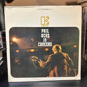May include: A square vinyl record jacket featuring a live performance photograph of a musician playing an acoustic guitar. The white cover displays the Elektra logo and the text Phil Ochs In Concert in a stylized font against a dark stage background.