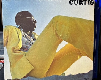 Può includere: Una copertina di un album di vinile vintage con un uomo in un abito giallo, seduto con le gambe incrociate, su uno sfondo di cielo blu. Il titolo dell'album "CURTIS" è stampato in lettere nere nell'angolo in alto a destra della copertina.