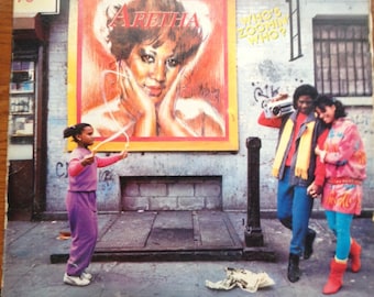 May include: A vintage vinyl record album cover featuring a portrait of Aretha Franklin with the album title "Who's Zoomin' Who?" in yellow text. The cover also features a young girl skipping rope in front of a brick building with a graffiti mural.