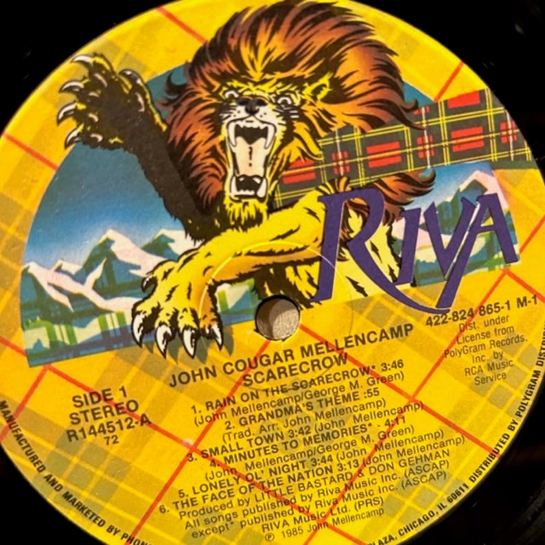 May include: A yellow vinyl record with a red and black plaid design. The record features a lion illustration and the text "John Cougar Mellencamp Scarecrow" and "Riva" in a purple font. The record is labeled "Side 1 Stereo R144512-A 72".