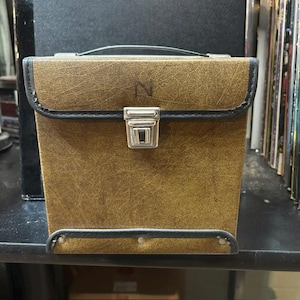 May include: A square-shaped storage case with a textured brown faux leather exterior and black trim. The front flap features a silver-toned metal latch closure and a small embossed letter N. A black handle is attached to the top of the case.