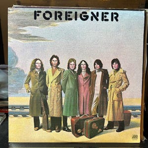 May include: A vintage vinyl record album cover featuring the band Foreigner. The cover art shows seven men in 1970s clothing standing in front of a train track. The album title "FOREIGNER" is printed in large, bold letters at the top of the cover.