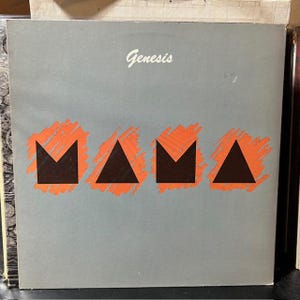 May include: A vinyl record album cover with the band name "Genesis" in white script at the top. The word "MAMA" is displayed in large, stylized black letters with orange accents. The background is a solid gray color.