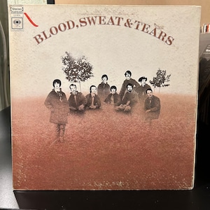 May include: A vintage vinyl record album cover featuring the band Blood, Sweat & Tears. The cover art shows the band members sitting in a field with trees in the background. The album title is printed in large, bold letters across the top of the cover.