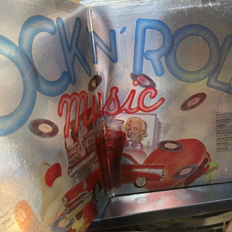 Pu&ograve; includere: Una copertina per disco in vinile apribile intitolata ROCK 'N' ROLL music con uno sfondo argento metallizzato. L'illustrazione presenta testo blu e rosso, un'auto d'epoca rossa, un hamburger, una ciliegia e un bicchiere di bibita rossa.
