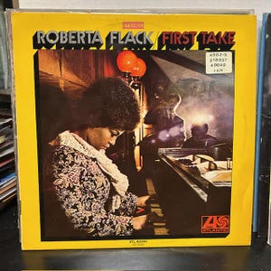 May include: A vintage vinyl record album cover for "Roberta Flack / First Take." The album cover is yellow with the artist's name and album title in bold black text. The image features Roberta Flack playing a piano, with other musicians in the background.