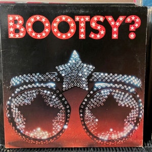 May include: A square vinyl record cover featuring the text BOOTSY? in white letters filled with red stars against a black background. Below the text is a graphic of star-shaped sunglasses covered in clear rhinestones on a red gradient base.