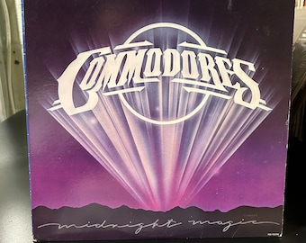 May include: A vintage vinyl record album cover featuring the band "Commodores" in a white, stylized font. The background is a purple gradient with a white circle and rays of light emanating from the center. The album title "Midnight Magic" is written in cursive at the bottom.