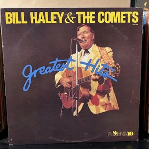 May include: A vinyl record album cover featuring Bill Haley & The Comets. The cover is brown with yellow text that reads "Bill Haley & The Comets" and "Greatest Hits". The album cover features a photo of Bill Haley playing a guitar. The album cover has a yellow star with the text "PHOENIX 10" in the bottom right corner.