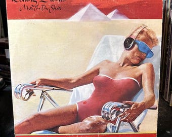 May include: A vinyl record cover for The Rolling Stones Made In The Shade featuring a person in a red swimsuit and blue visor sitting in a lounge chair. The background shows white pyramids under a red sky with the band name and album title text.
