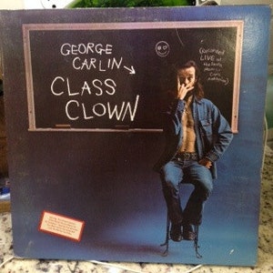 May include: Album cover for George Carlin's "Class Clown." The cover features a chalkboard with the title and artist's name in white chalk. Carlin is seated, wearing a denim shirt and jeans. The album is labeled "(Recorded LIVE at The Santa Monica Civic Auditorium)."
