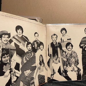 May include: A black and white photo of a band with 9 members. The band members are all wearing casual clothing and are standing in front of a white background. The photo is from the 1970s.