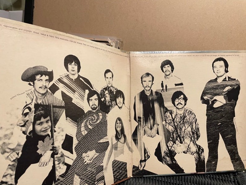 May include: A black and white photo of a band with 9 members. The band members are all wearing casual clothing and are standing in front of a white background. The photo is from the 1970s.
