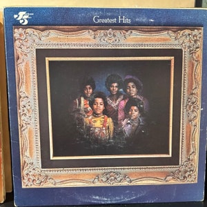 May include: A square vinyl record cover featuring a group portrait of five individuals in a dark setting, framed by an ornate, weathered gold-toned border. The top of the blue cover displays the text Greatest Hits and a small logo with the number 5.