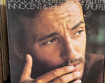 May include: A vintage vinyl record album cover featuring a close-up of Bruce Springsteen's face. The album title is "Bruce Springsteen: The Wild, The Innocent & The E Street Shuffle".