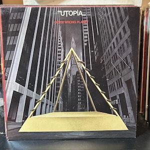 May include: A vinyl record album cover for Utopia's "Oops! Wrong Planet." The cover features a black and white cityscape with a gold-colored geometric structure in the foreground. The band's name and album title are printed at the top.