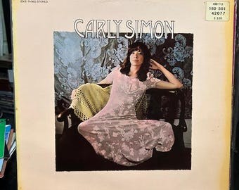 May include: Album cover featuring Carly Simon in a pink floral dress, seated in a dark chair. The background has a floral pattern. The album title "CARLY SIMON" is displayed in large letters. The album cover has the text "42077 (EKS-74002) STEREO" and "498 1-3 180 581 42077 $2.50".