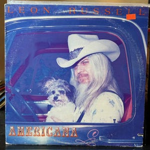 May include: A vintage vinyl record album cover featuring Leon Russell. The cover art shows Leon Russell in a cowboy hat, holding a small dog, with the album title "Americana" in red letters.