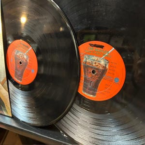 Pu&ograve; includere: Due dischi in vinile neri con etichette centrali arancioni brillanti. Ogni etichetta mostra la grafica di un bicchiere riempito con una bevanda scura e una cannuccia, insieme al testo The Beatles Rock 'N' Roll Music e varie tracce.