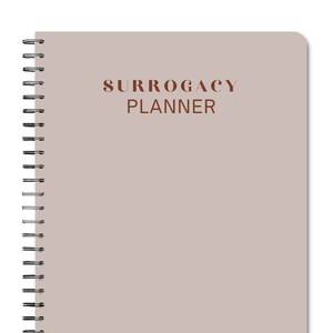 May include: A beige spiral bound notebook with the text "SURROGACY PLANNER" printed in brown on the front cover.