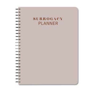 May include: A light gray spiral bound notebook with the text "SURROGACY PLANNER" printed in brown on the front cover.