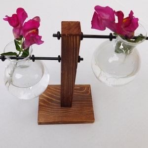 May include: A wooden stand with two glass bulbs for holding flowers. The stand is brown and has a black metal frame. The bulbs are clear and filled with water. Pink flowers are in each bulb.
